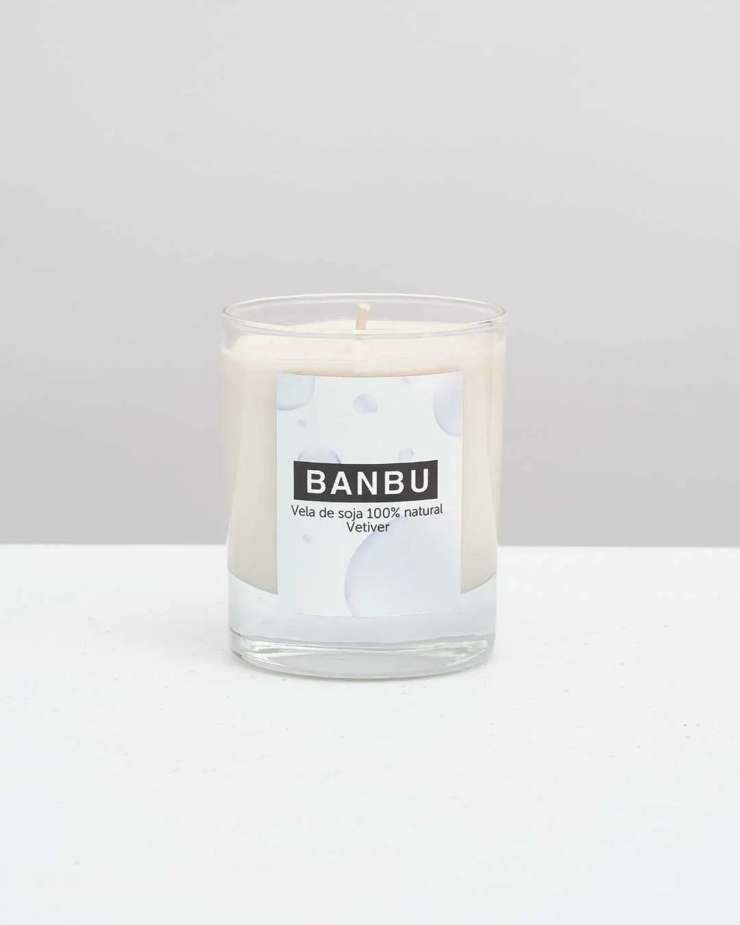 Vela de soja Banbu con aroma a vetiver en vaso de cristal transparente, sobre superficie blanca y fondo neutro.