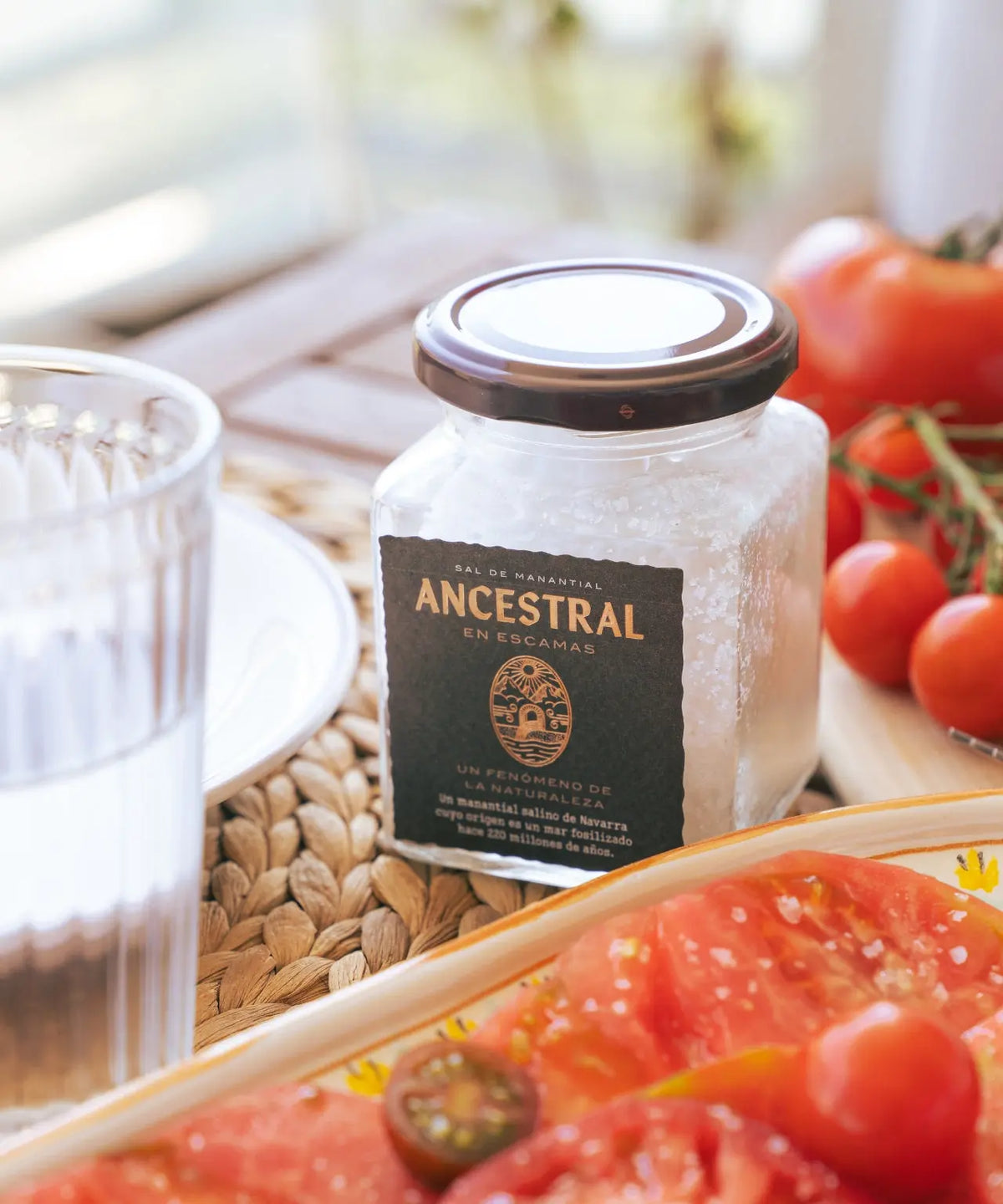 Glass jar of natural spring salt flakes with a black label on a wicker mat next to fresh tomatoes.