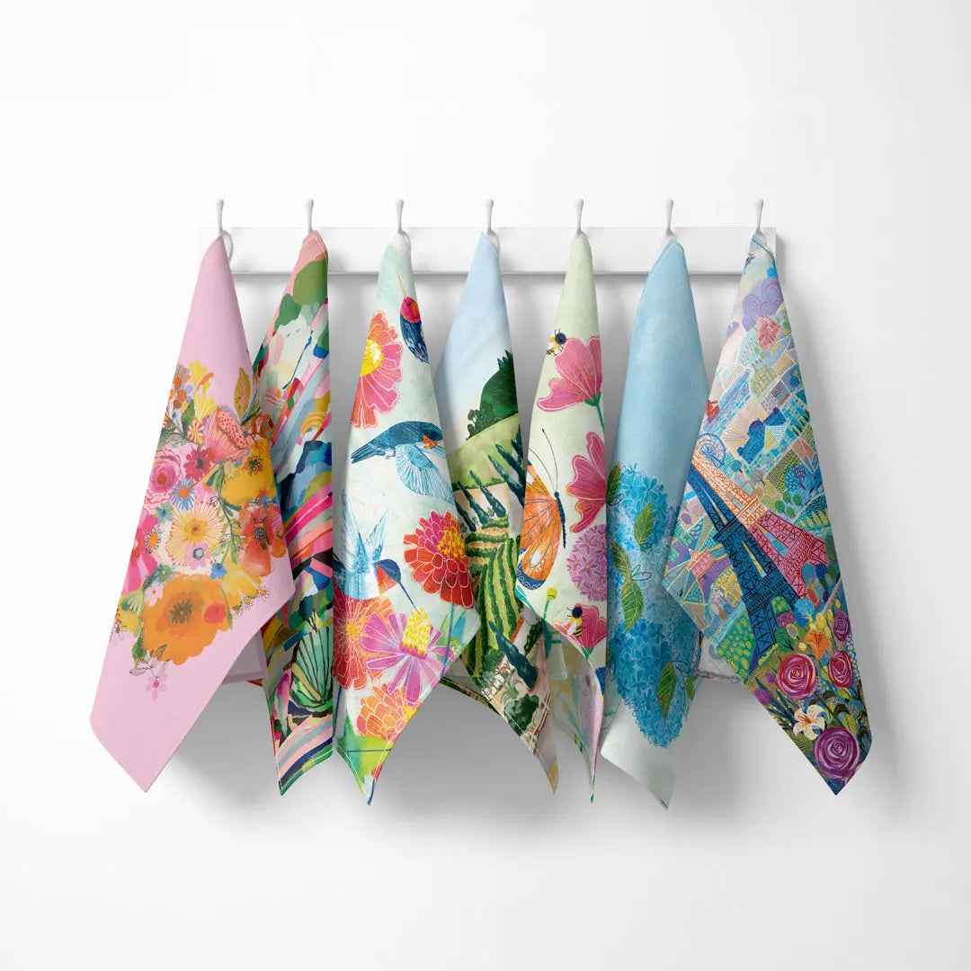 Seven colorful, artistic cotton tea towels featuring floral, nature, and Paris Eiffel Tower designs, hanging on a rack.