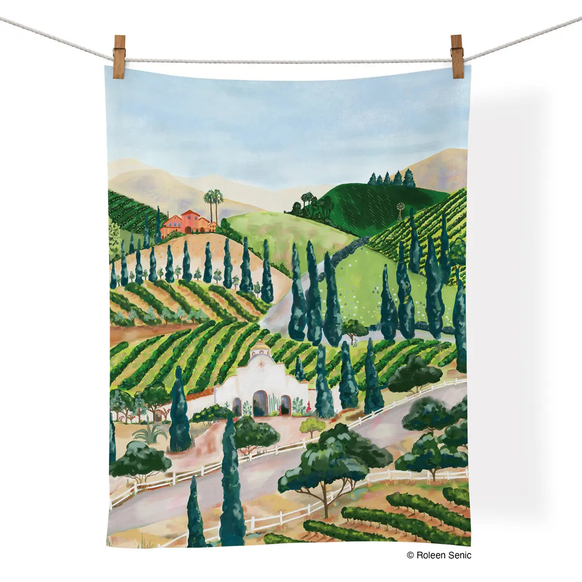 Cotton tea towel hanging on a line, featuring a painted landscape of rolling vineyards, cypress trees, and buildings.