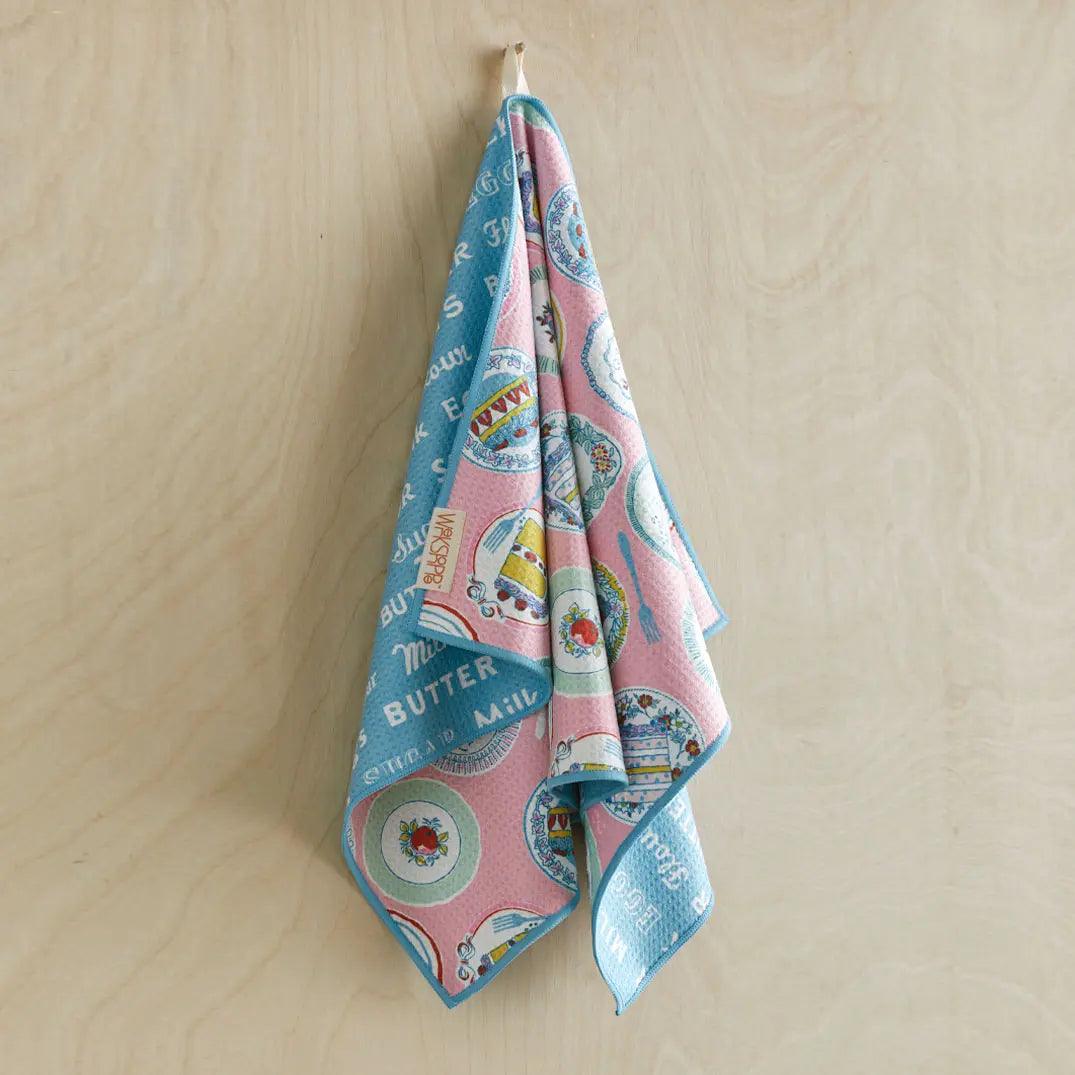 Microfiber kitchen towel in pink and blue featuring a cake and plate pattern, hanging against a light wood background.