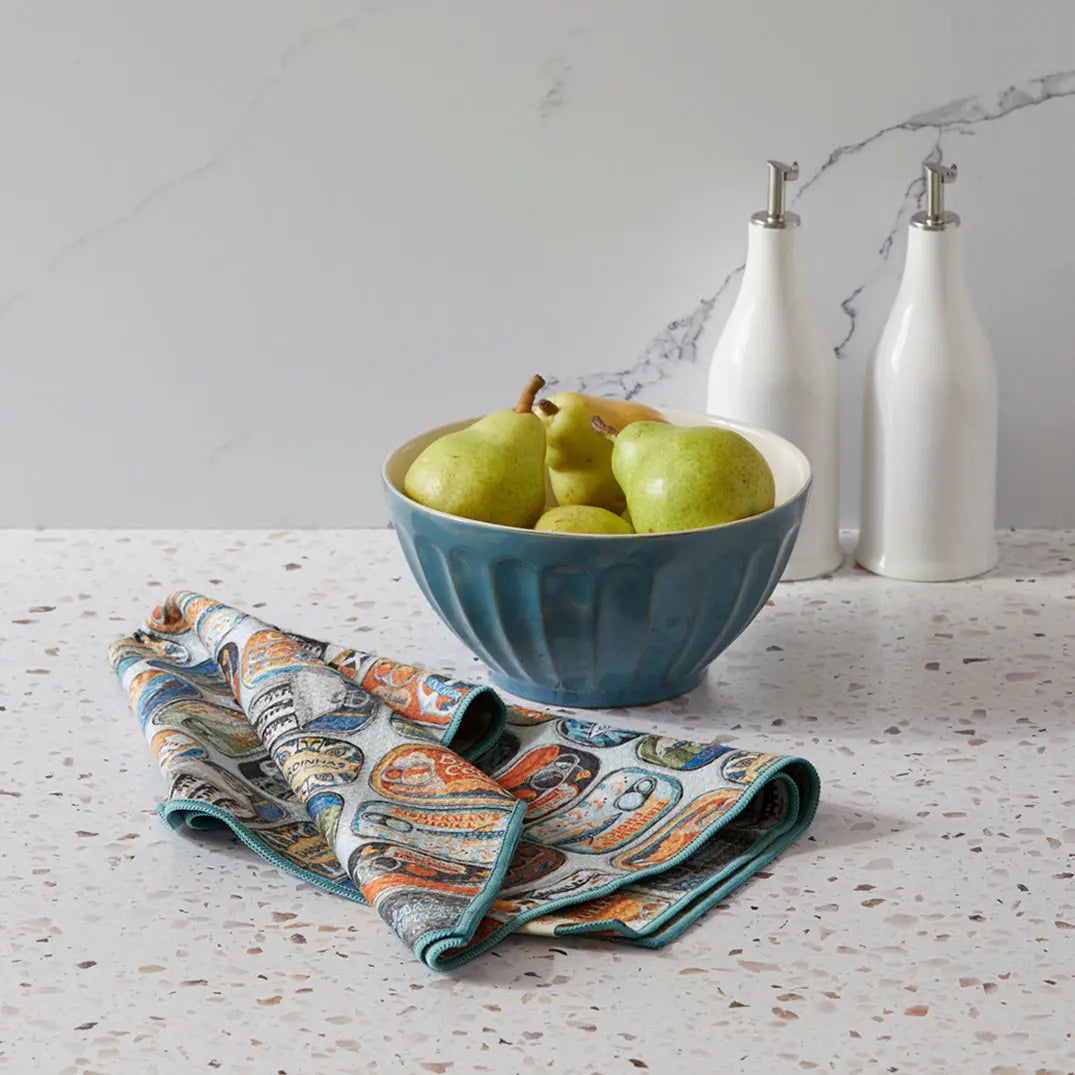 Microfiber towel featuring a sardine tin print in orange and blue, folded on a speckled countertop near a pear bowl.