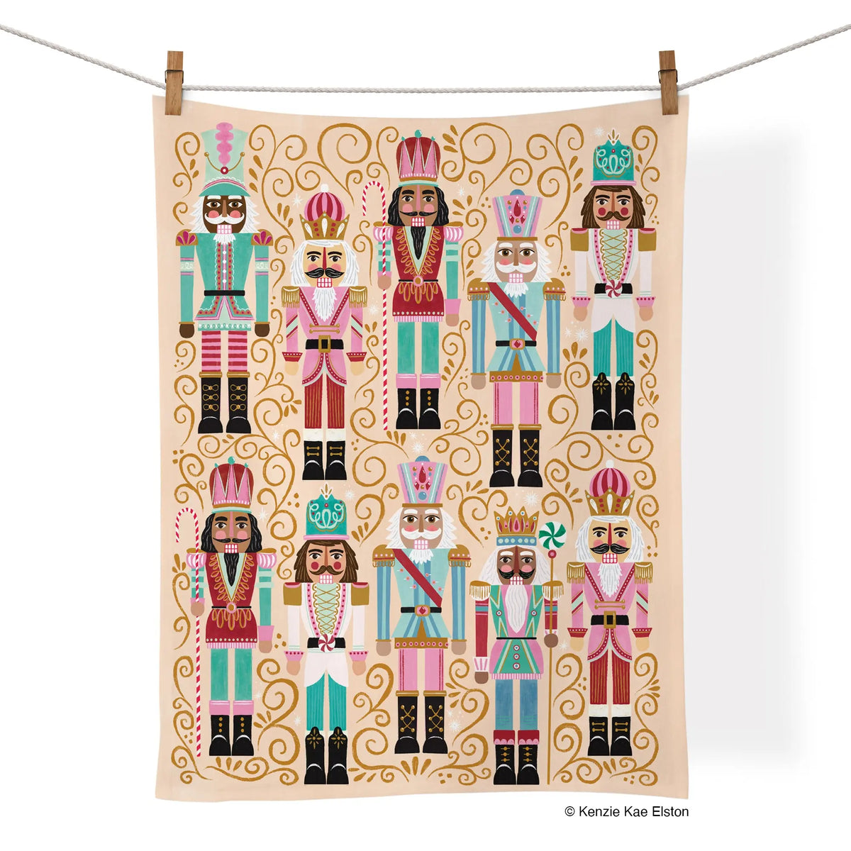 Cotton tea towel with festive nutcracker illustrations in pink, teal, and gold on a soft peach background.