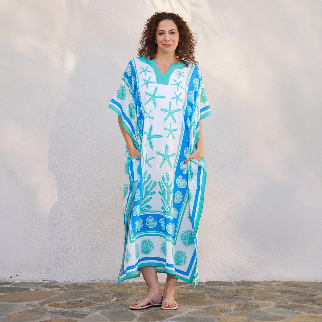 Woman in a white caftan with blue starfish and seashell prints, V-neck, and side pockets.