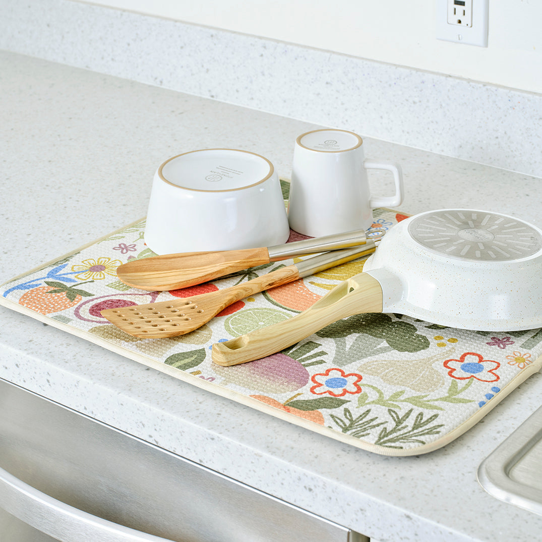 Rectangular fabric dish drying mat with a colorful, whimsical fruit and floral pattern, holding kitchenware on a counter.