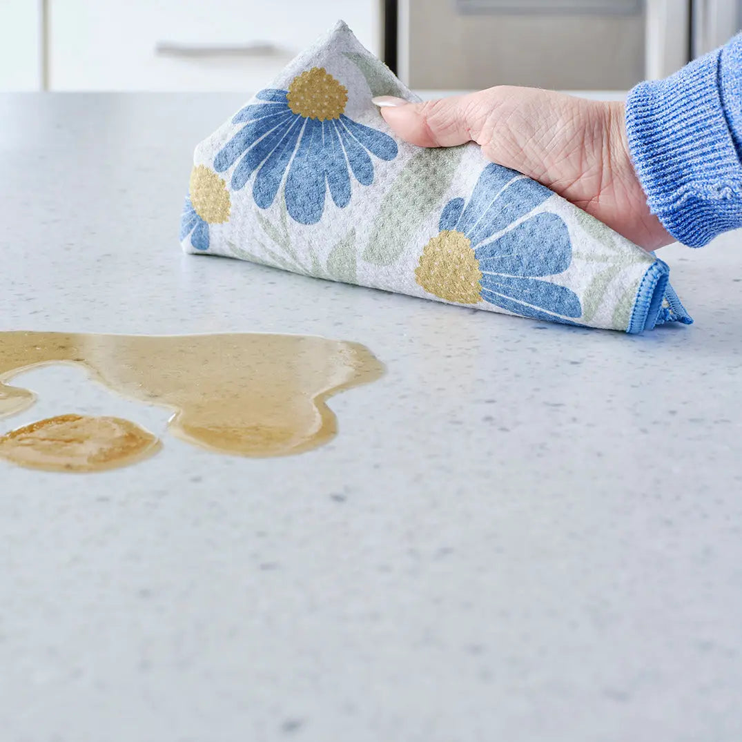 Hand cleaning a spill on a countertop with a white towel featuring a blue and yellow coneflower floral pattern.