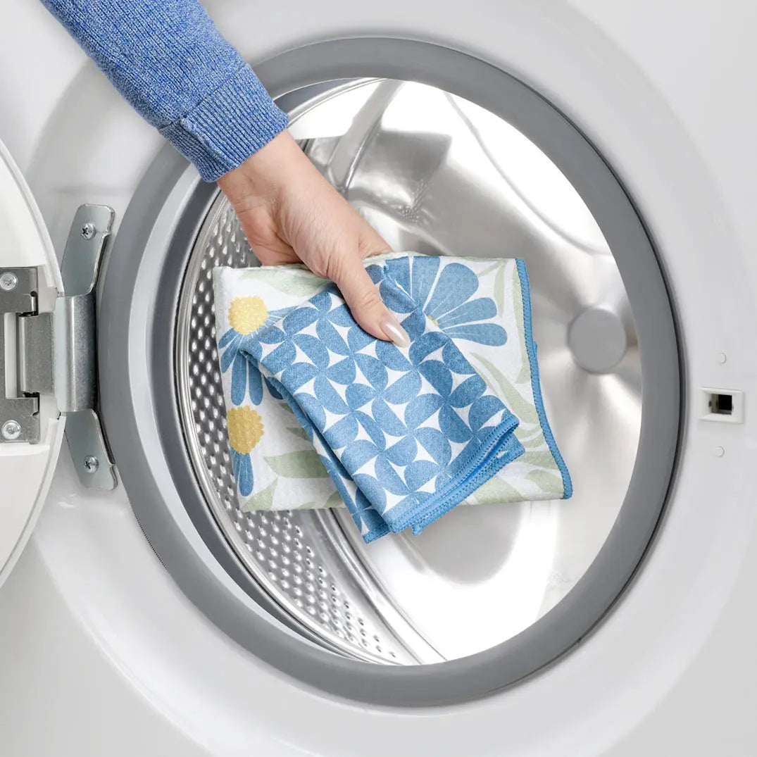 A hand places folded blue and yellow coneflower-patterned towels into an open washing machine drum.