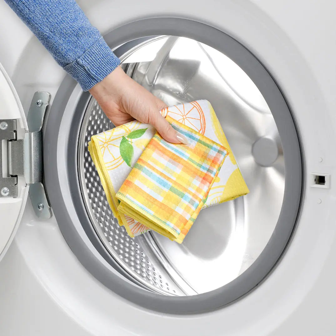 Hand placing yellow lemon-patterned and plaid-checkered towels into a white washing machine.