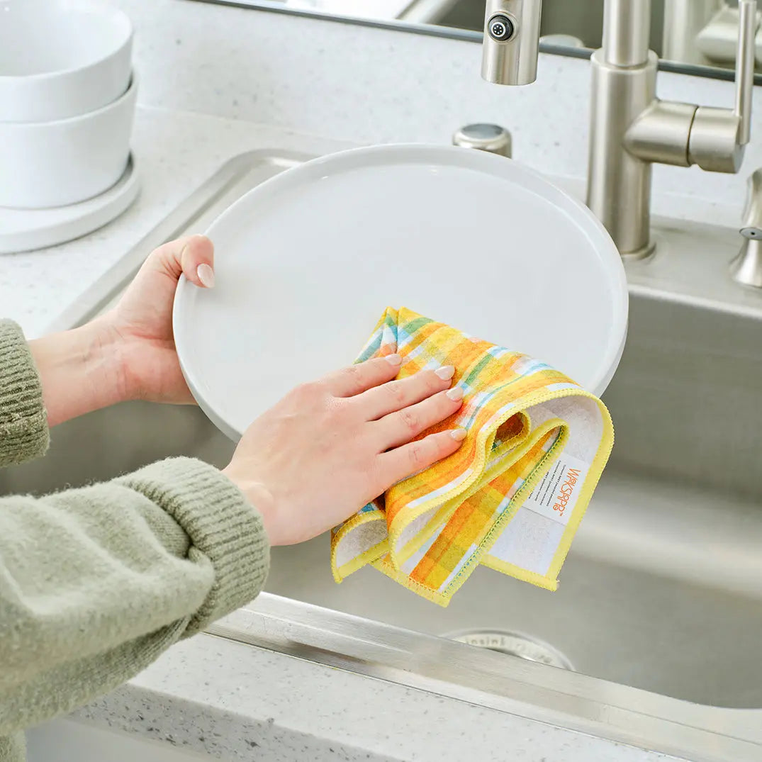 Hands drying a white plate with a yellow and blue plaid patterned fabric towel over a kitchen sink.