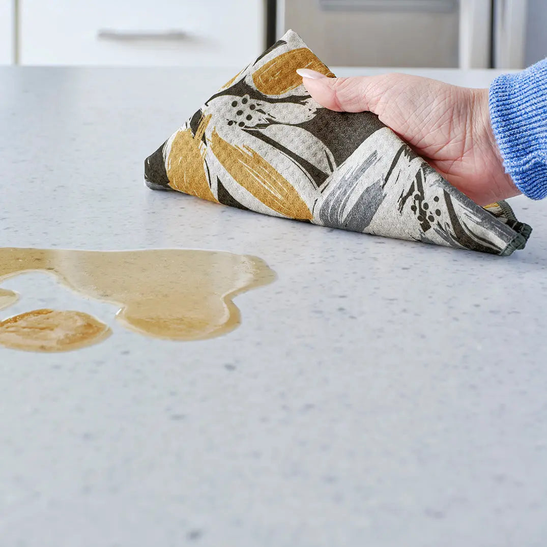 Hand wiping a spill on a countertop with a soft, absorbent towel featuring an abstract yellow and gray leaf pattern.