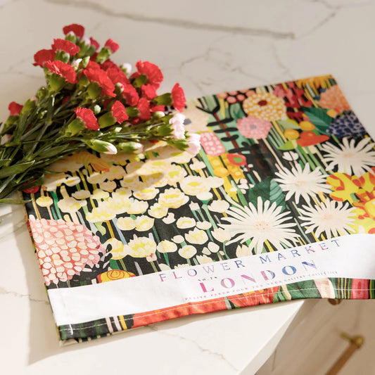 Cotton tea towel with vibrant floral print on black background and "Flower Market London" text, beside red flowers.