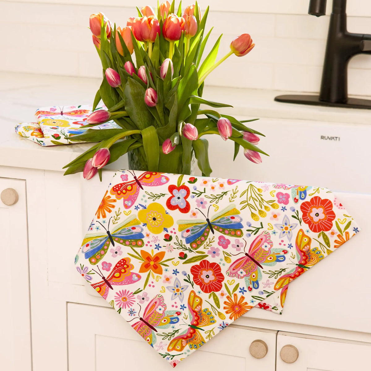 Cotton tea towel with vibrant butterfly and floral print in red, yellow, blue, and pink, hanging on a white sink.