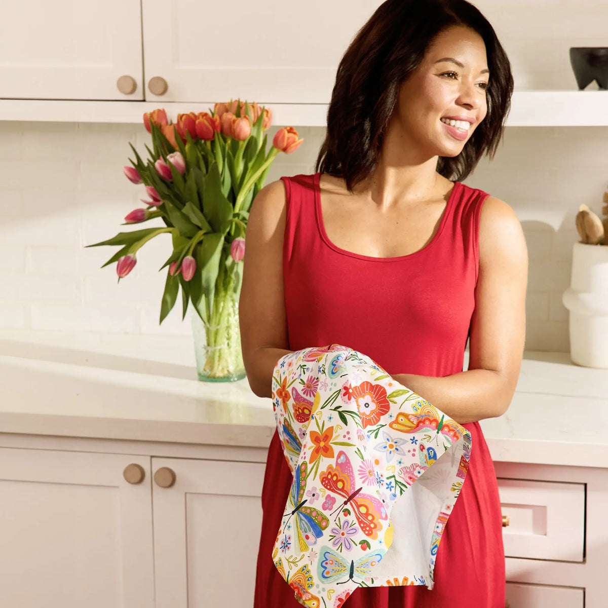 A woman in a red dress holds a white cotton tea towel featuring a colorful butterfly and floral print pattern.