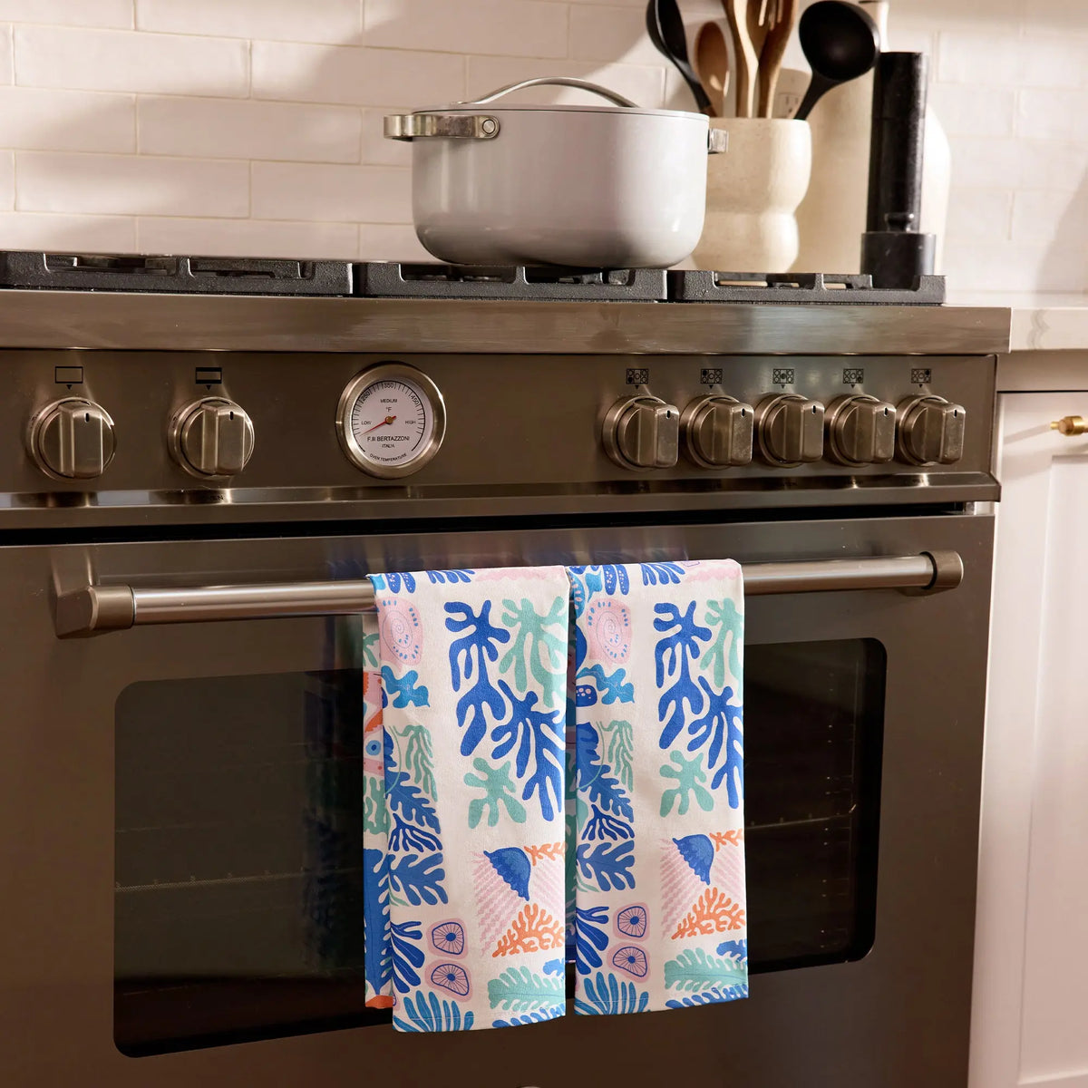 Cotton tea towel with blue, pink, and orange coastal coral patterns draped over a modern stainless steel oven handle.