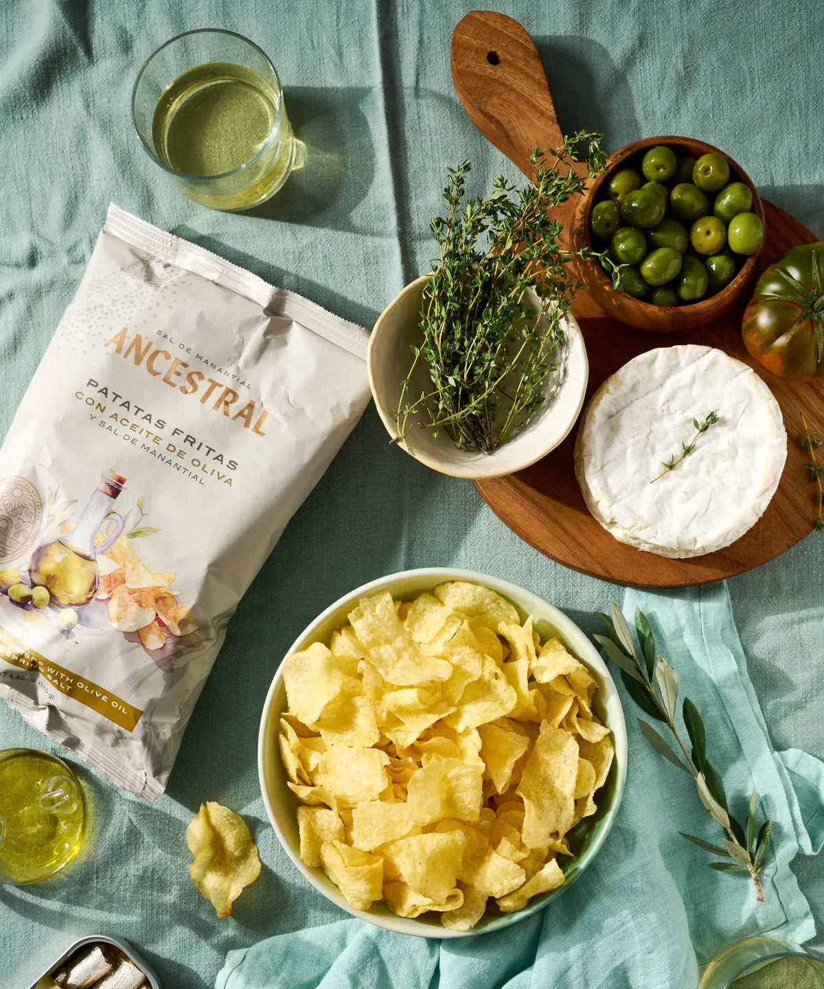Bag of gourmet olive oil potato chips next to a bowl of chips, fresh thyme, olives, camembert, and a wooden board.