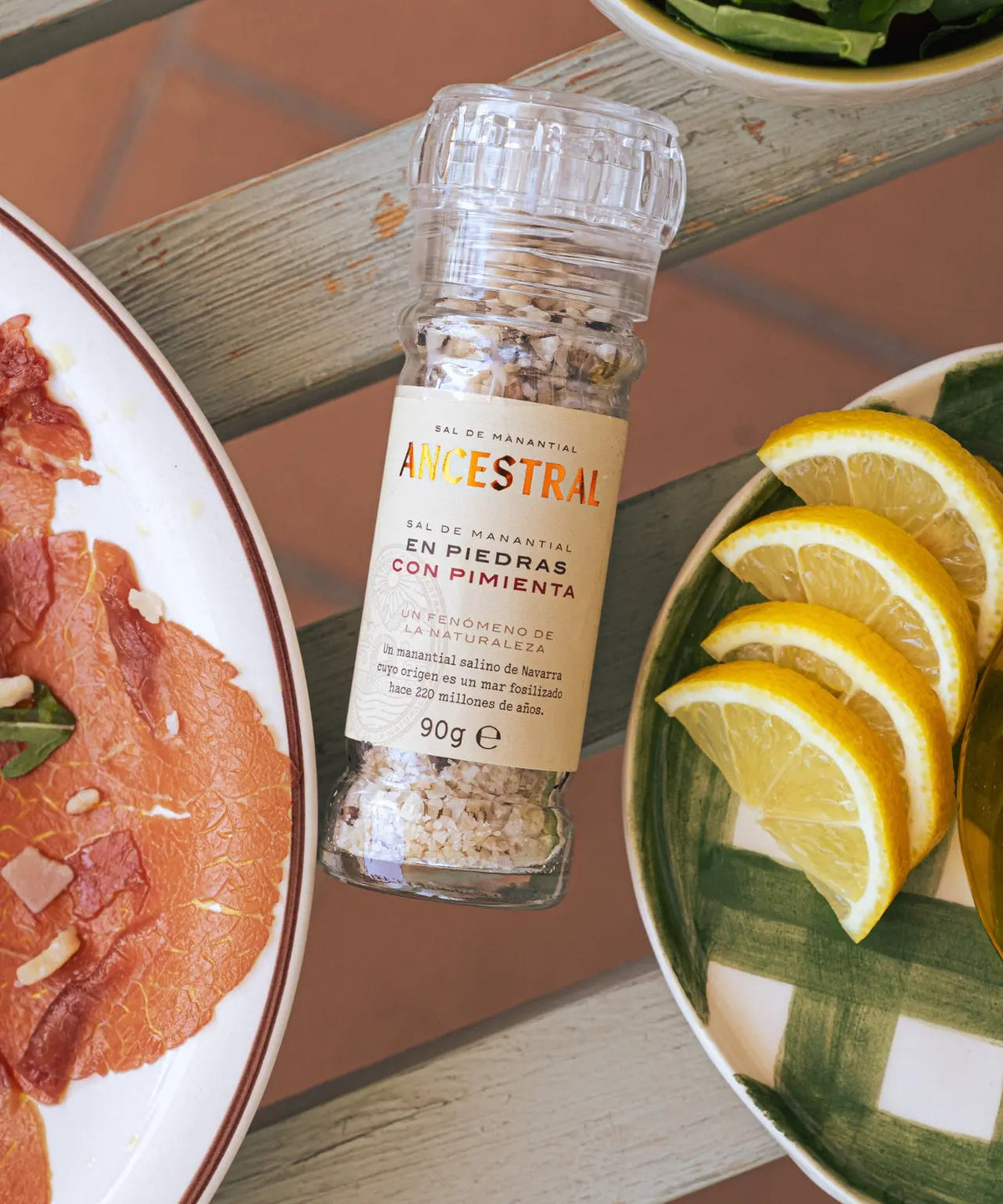 A glass salt grinder with coarse salt and pepper on a wooden table, next to plates with sliced meat and lemon.