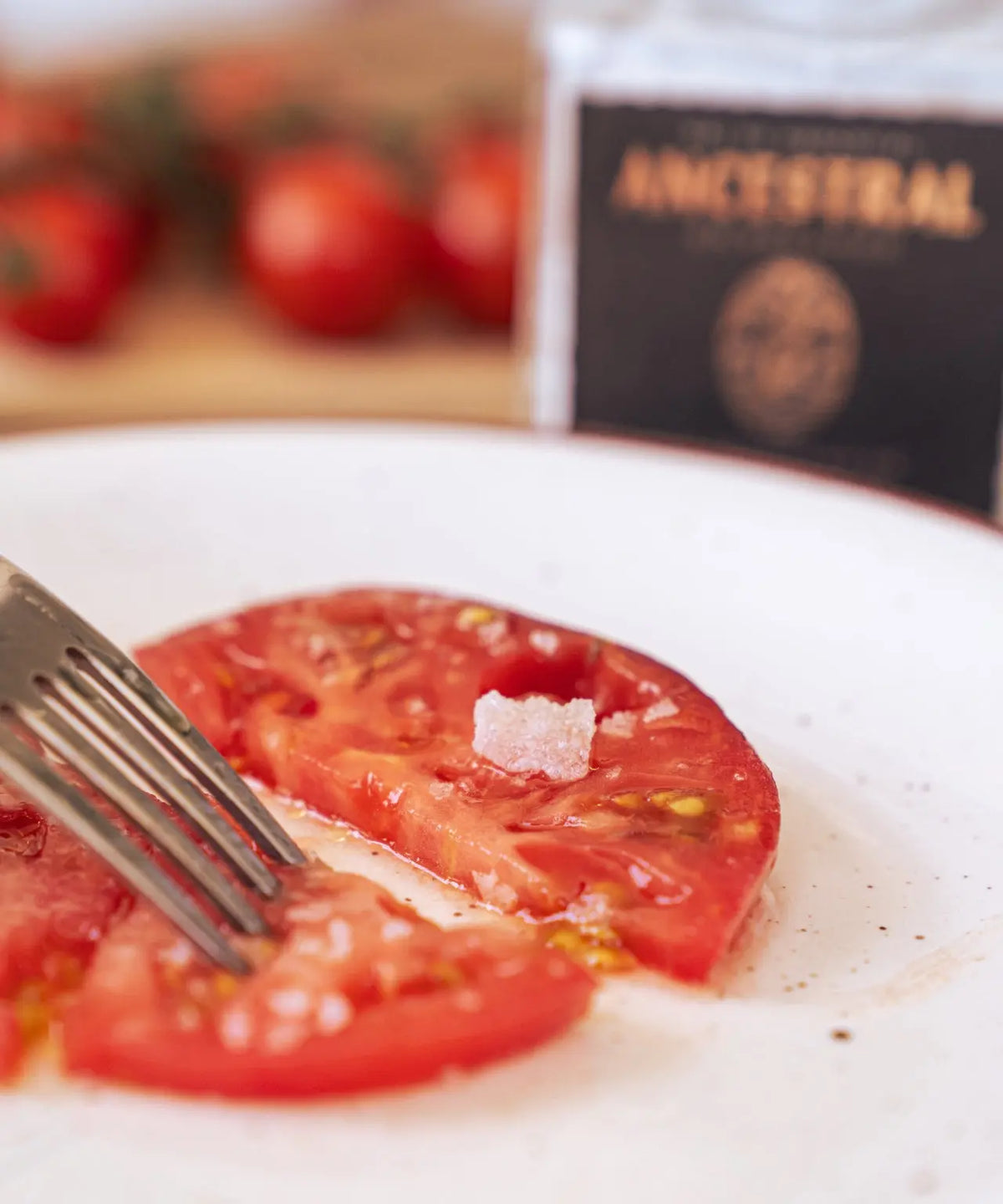 Sliced red tomato on a white plate with a metal fork and coarse natural salt flakes, blurred background.