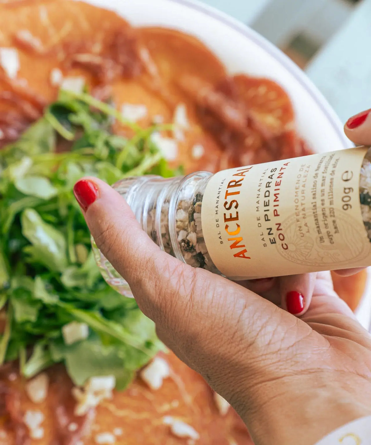 Hand with red nail polish holding a clear salt grinder over a plate of carpaccio with fresh greens and cheese.