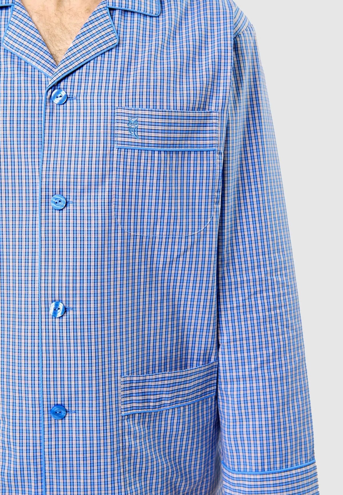 Chaqueta de pijama azul a cuadros con solapa, botones a tono, bolsillo en pecho con logo de búho y vivo decorativo.