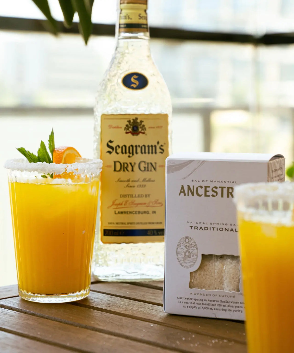 Natural spring salt box next to a gin bottle and two orange cocktails with salted rims on a wooden table.