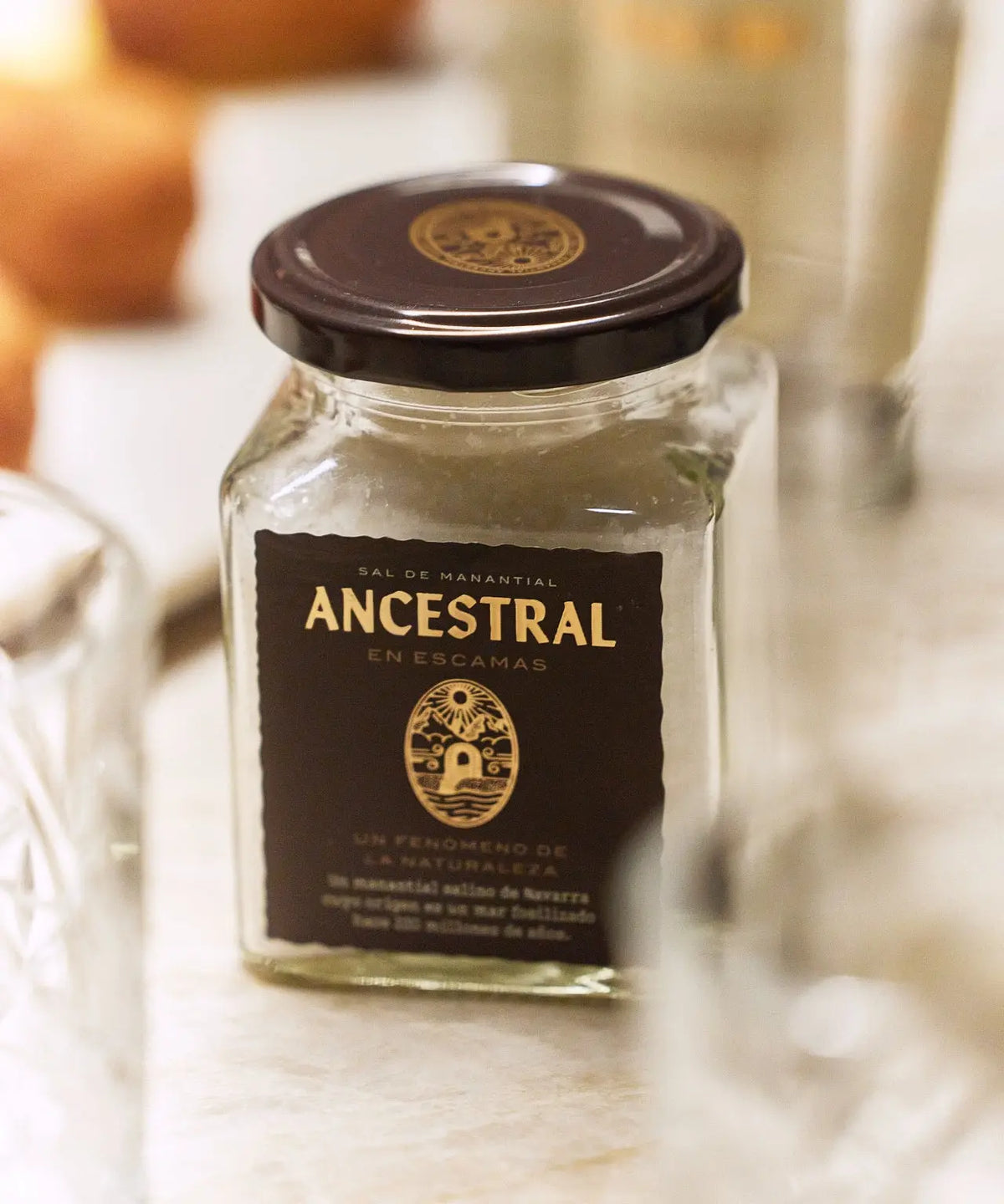 Square glass jar of spring salt flakes with a dark brown label and black lid, set on a bright, blurry background.