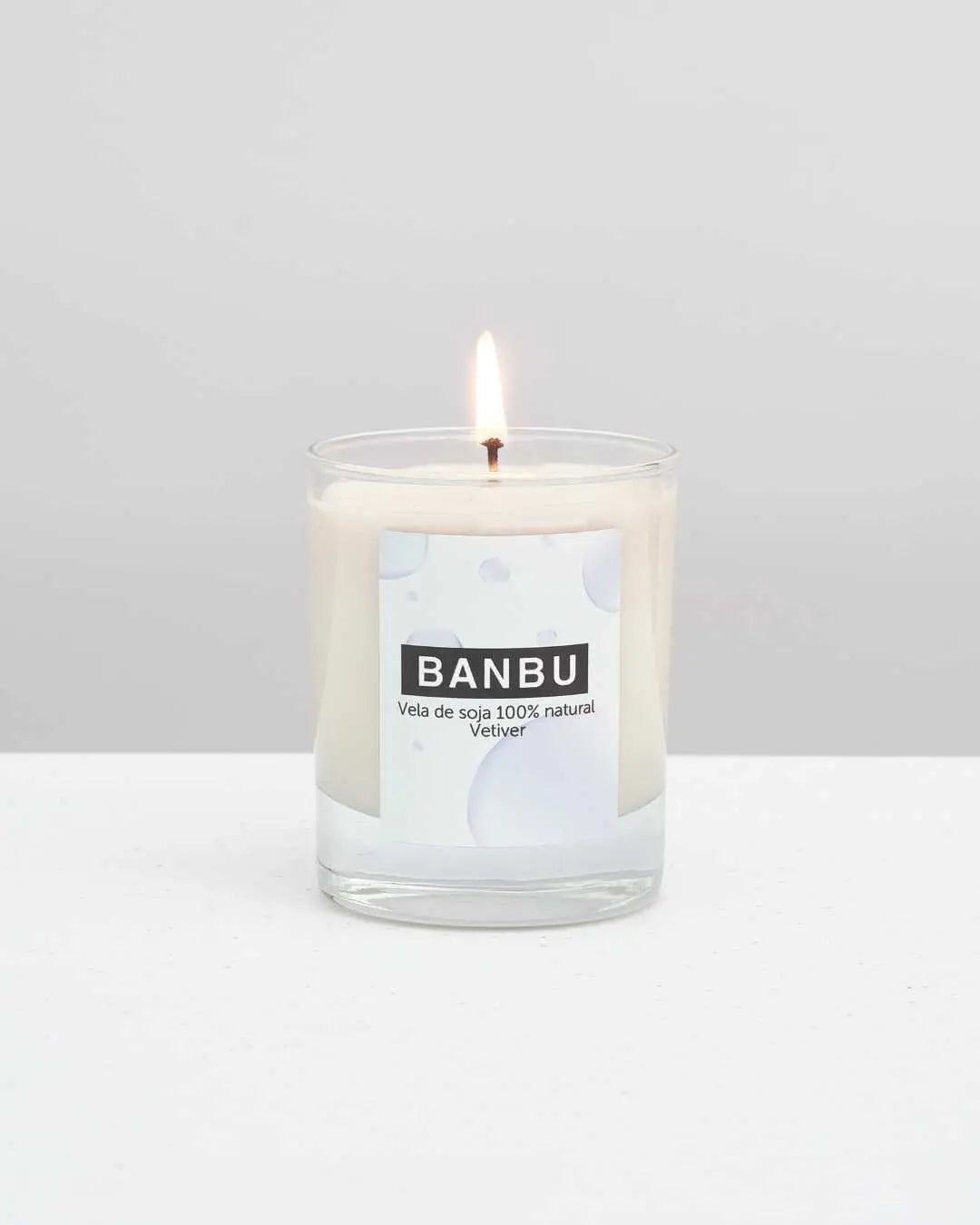 Vela de soja encendida marca Banbu, aroma Vetiver, en vaso de cristal sobre fondo neutro y superficie blanca.