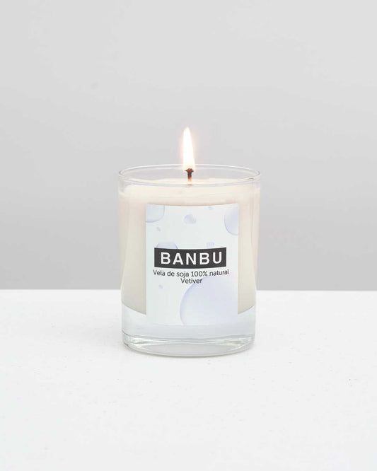 Vela de soja encendida marca Banbu, aroma Vetiver, en vaso de cristal sobre fondo neutro y superficie blanca.