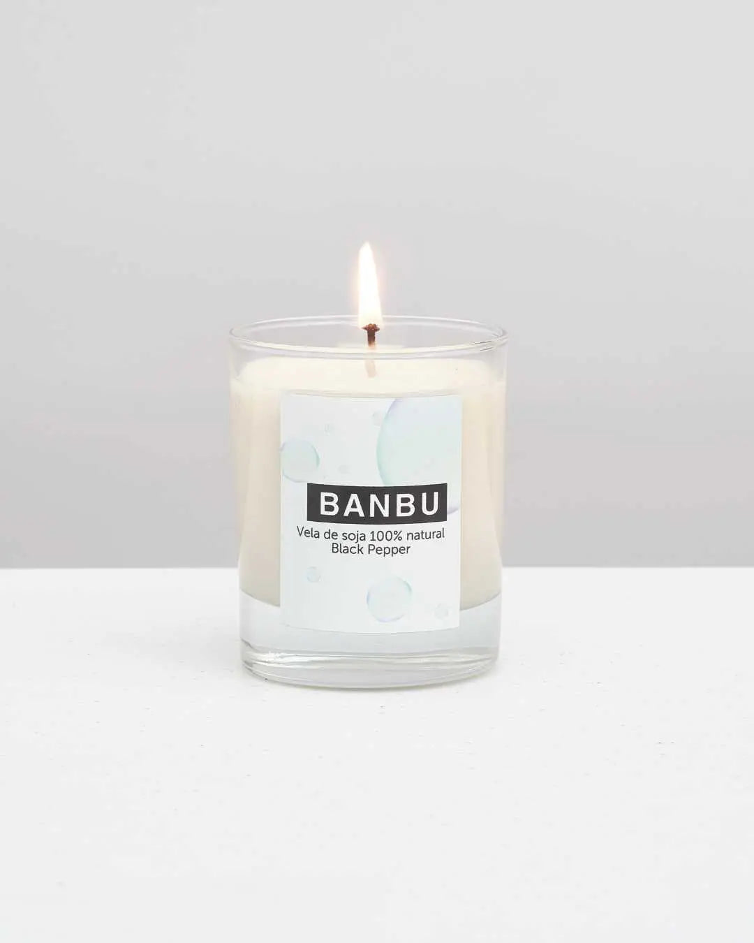 Vela de soja natural BANBU Black Pepper encendida en vaso de vidrio sobre superficie blanca. Diseño minimalista.