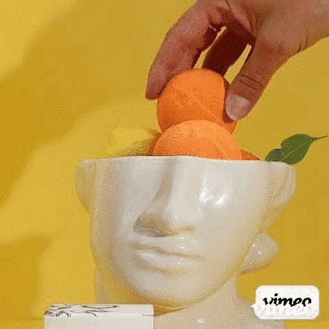Mano colocando bombas de baño naranjas de Vitamina C en un recipiente de cerámica con forma de cabeza sobre fondo amarillo.