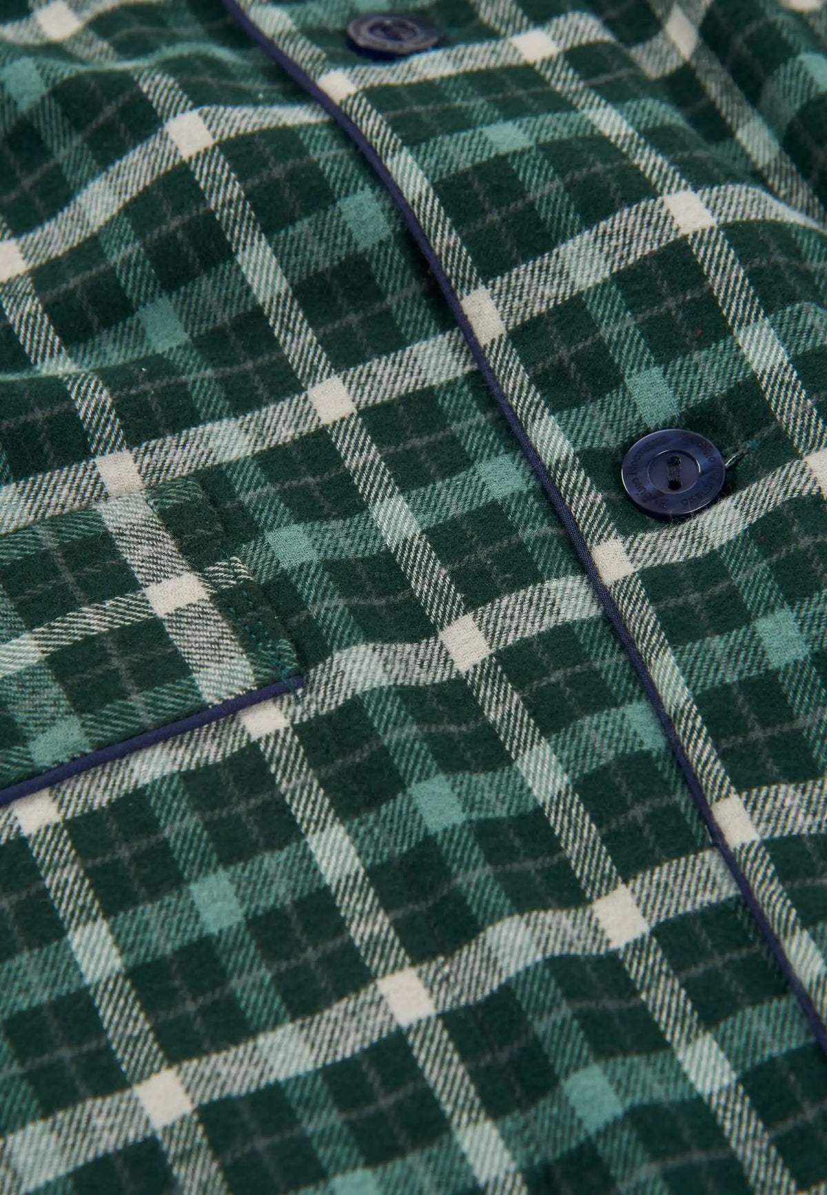 Detalle de pijama de franela verde a cuadros con botones azul marino y bolsillo frontal, tejido suave y texturizado.