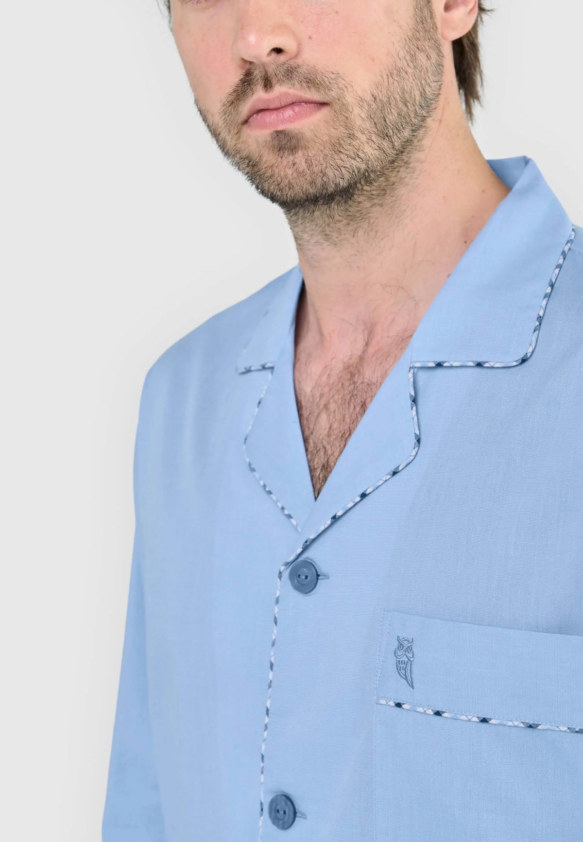 Pijama azul celeste de hombre con solapas, ribete de cuadros, botones frontales y bolsillo con logo bordado.