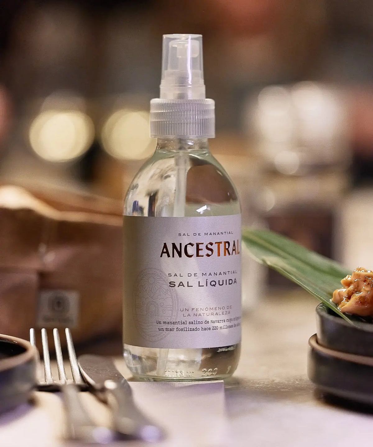Clear 200ml spray bottle of liquid spring salt with a white label, positioned on a blurred restaurant dining table.