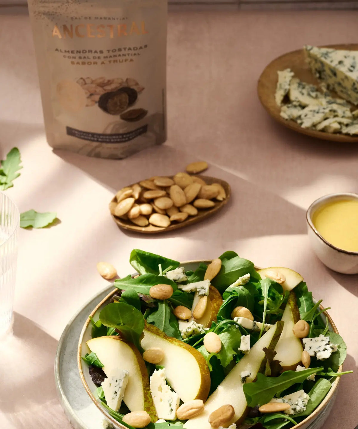 Truffle-salted roasted almonds next to a salad bowl with pear, blue cheese, and greens on a light pink surface.