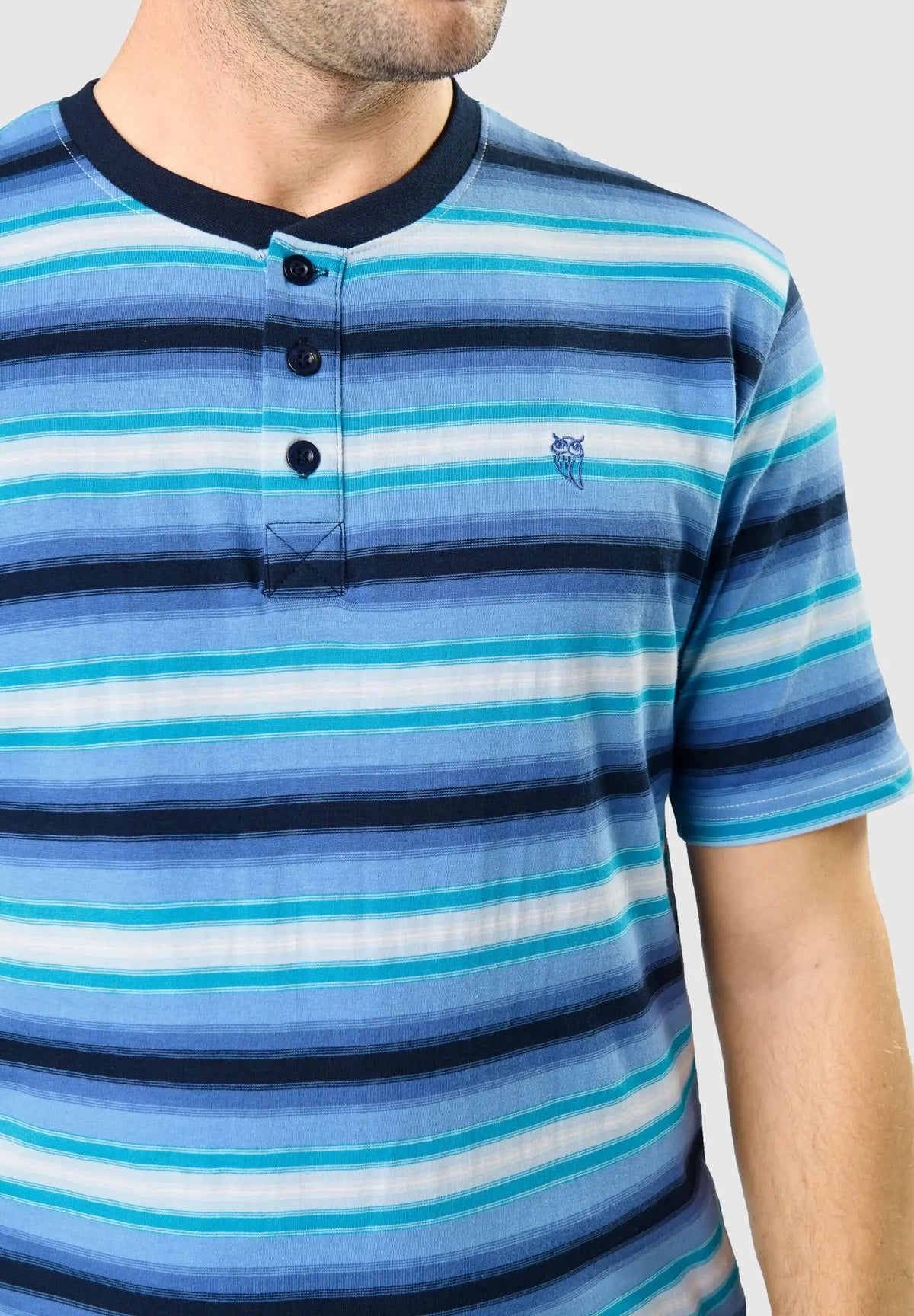Camiseta de pijama a rayas azules, cuello redondo con tres botones y logo de búho bordado, puesta por un hombre.