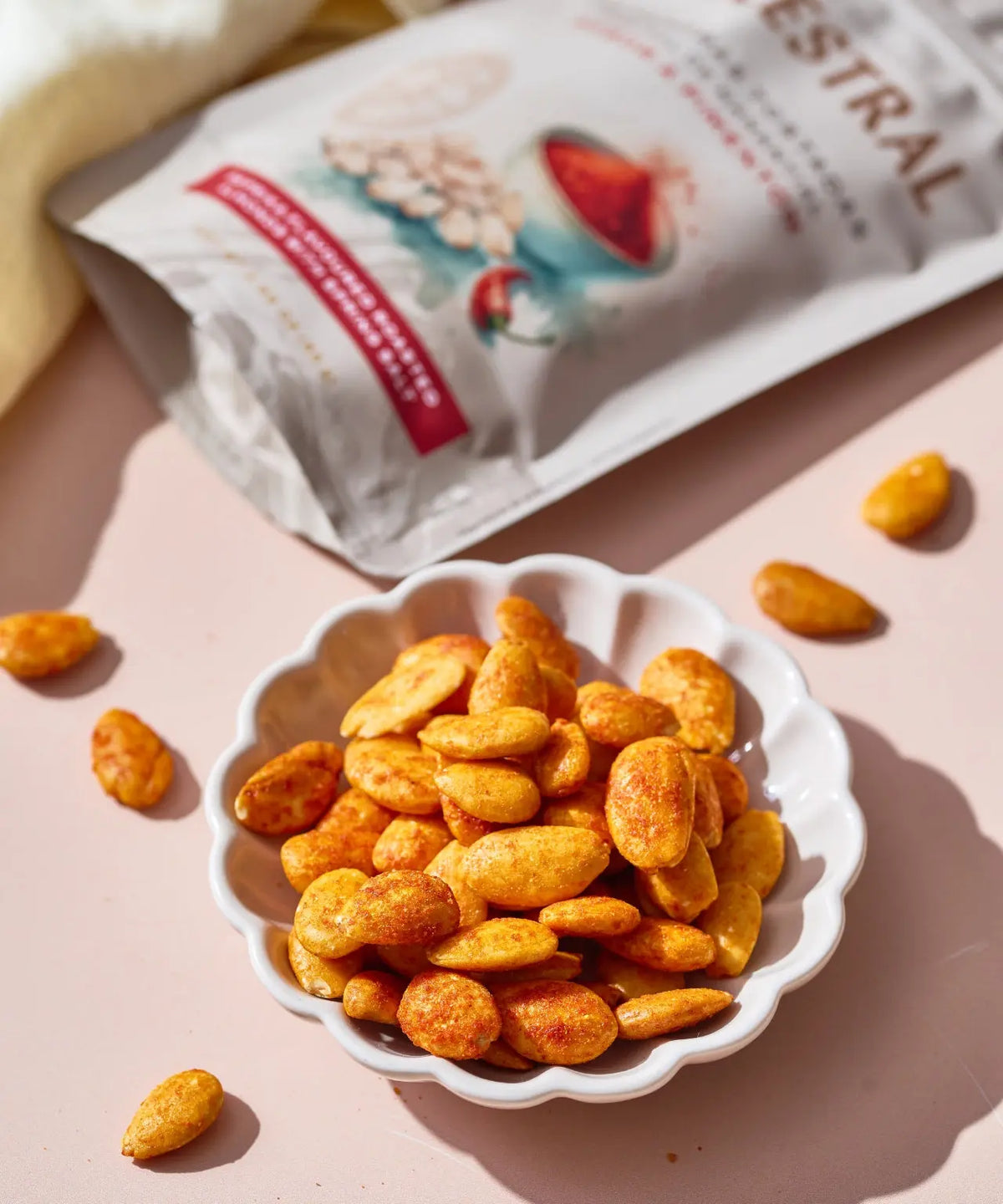 Roasted almonds seasoned with paprika in a white scalloped bowl, next to a branded snack pouch on a pink surface.