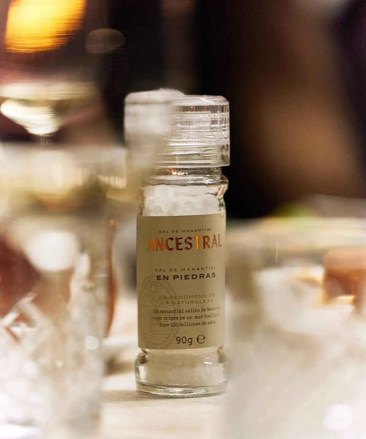 Clear glass mill filled with natural spring salt crystals, featuring a tan label with text, centered on a table.