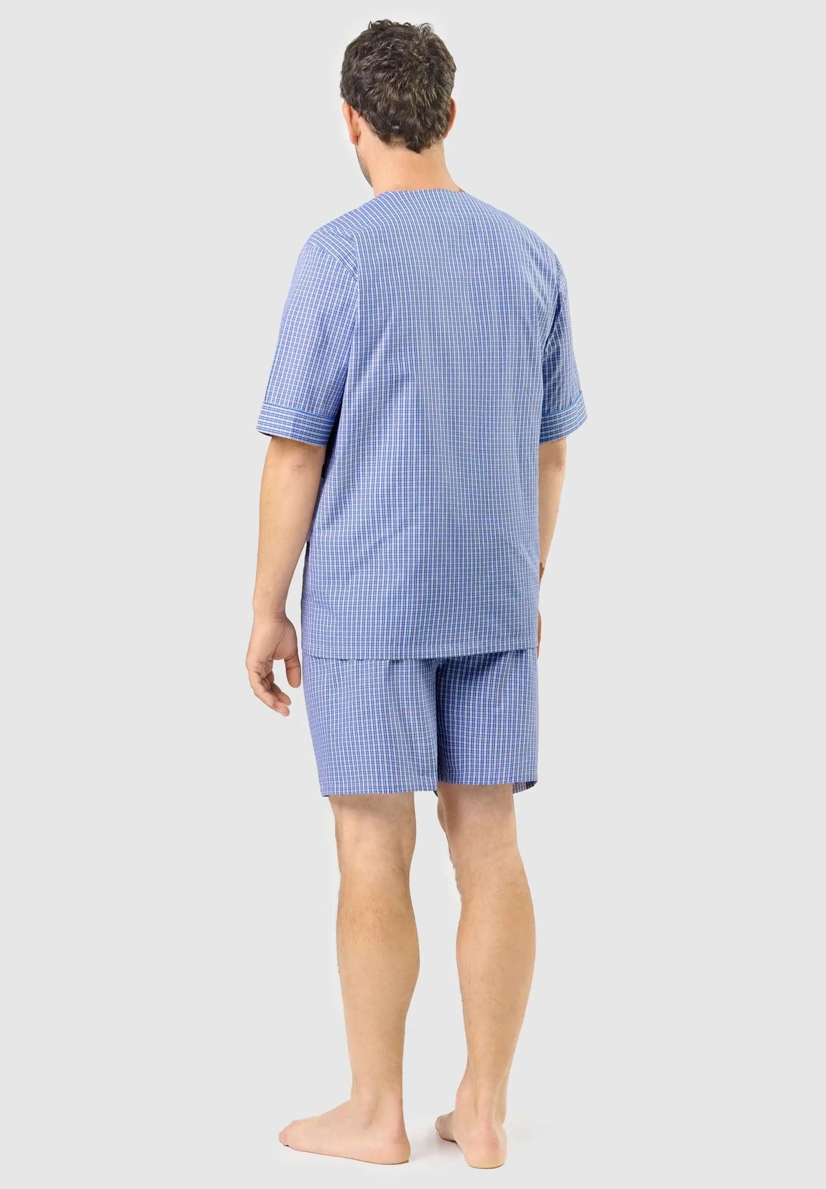 Hombre de espaldas con pijama corto de verano azul a cuadros pequeños, camiseta manga corta y pantalón, fondo gris.