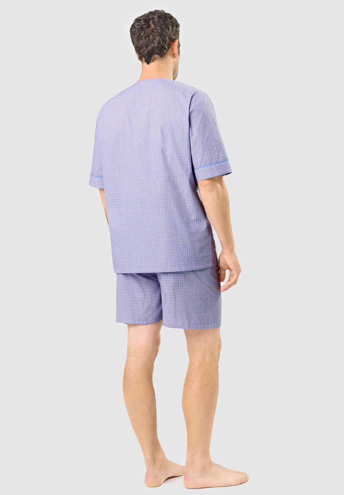 Hombre viste pijama corto de algodón azul con estampado de cuadros finos y ribete azul en las mangas. Vista trasera.