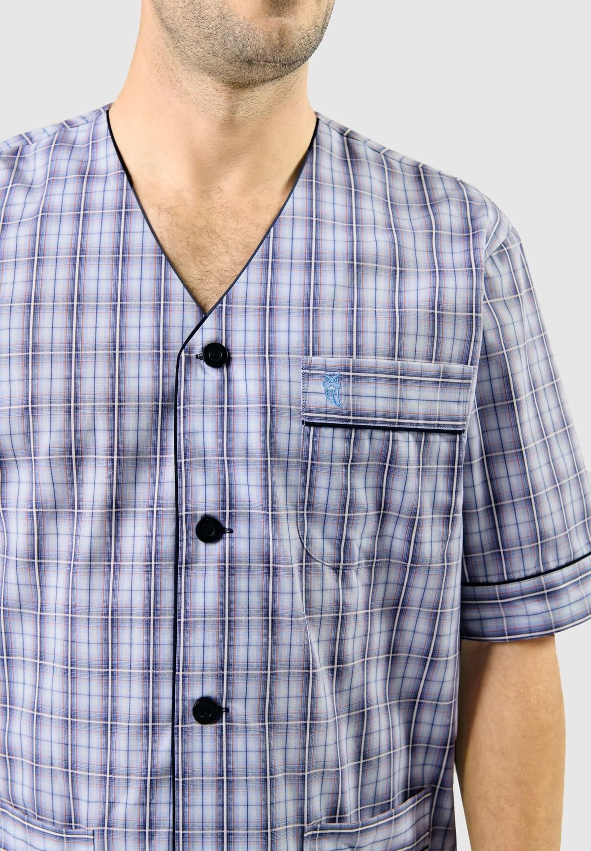 Pijama de hombre azul a cuadros, cuello pico, botones oscuros, bolsillo con logo de búho y manga corta sobre modelo.