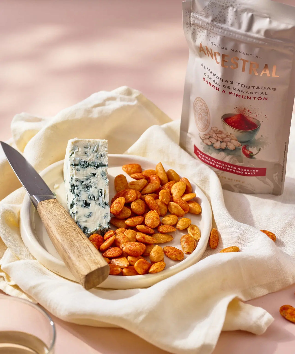Roasted paprika almonds and a slice of blue cheese on a plate with a wooden knife, beside an Ancestral snack pouch.