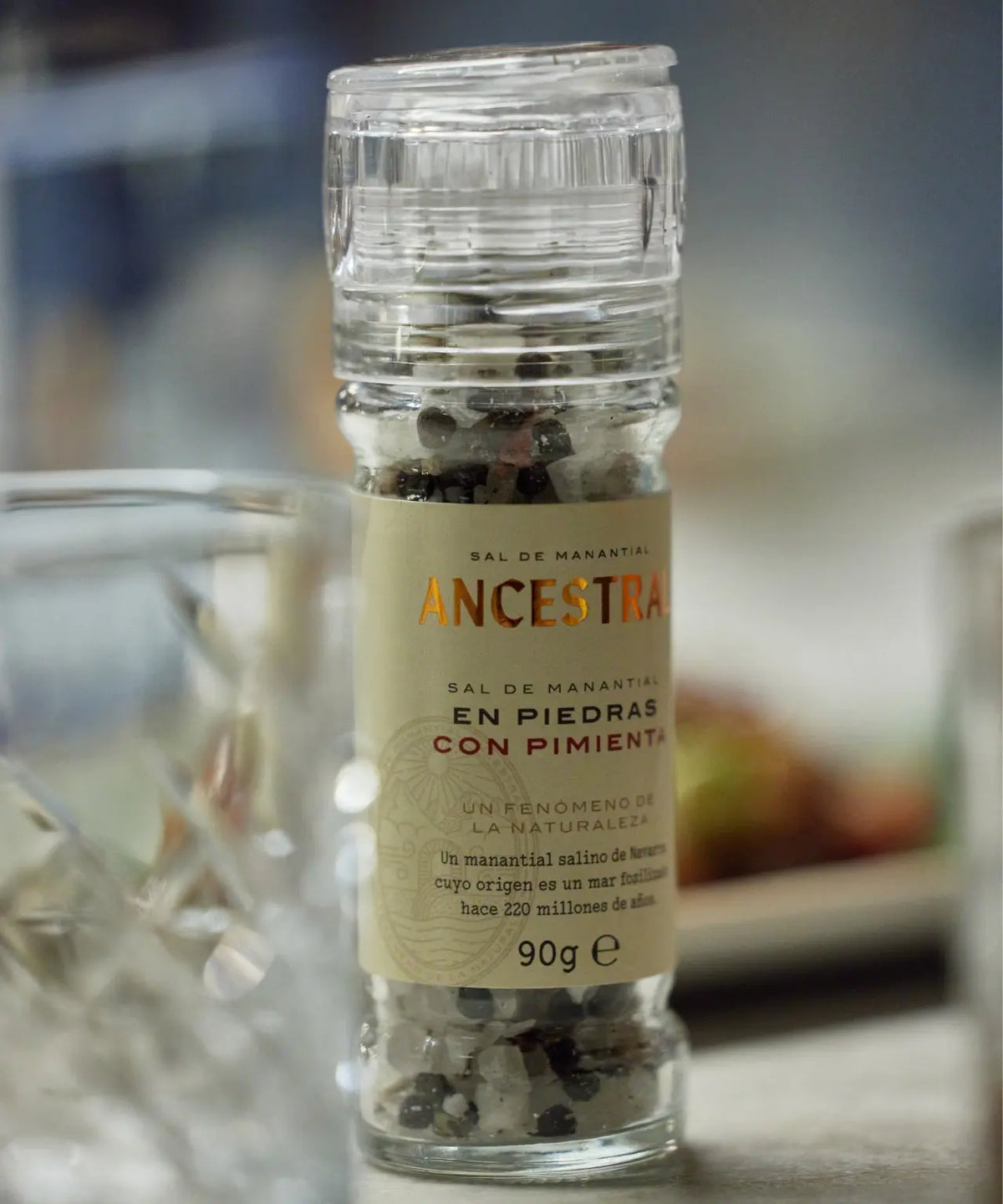Clear glass grinder filled with salt crystals and four-pepper mix, featuring a beige label with Ancestral branding.