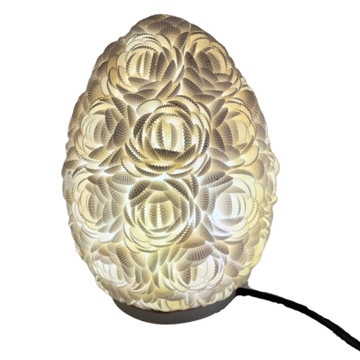 Lámpara boho ovalada de 20cm, hecha con pequeñas conchas blancas dispuestas en forma de rosas, emitiendo luz cálida.