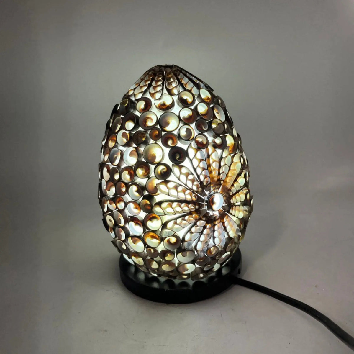 Lámpara boho ovalada de 20 cm, hecha de conchas marinas color chocolate con diseño de flor iluminada sobre base negra.