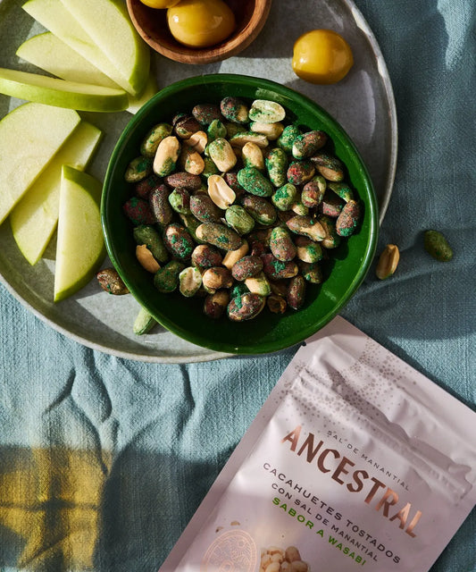 A bowl of green wasabi-coated roasted peanuts served with fresh green apple slices on a blue linen tablecloth.