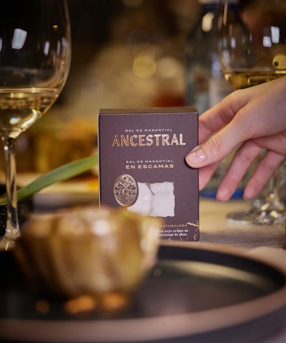 A hand holds a dark brown box of gourmet spring salt flakes with a clear window revealing white salt crystals.