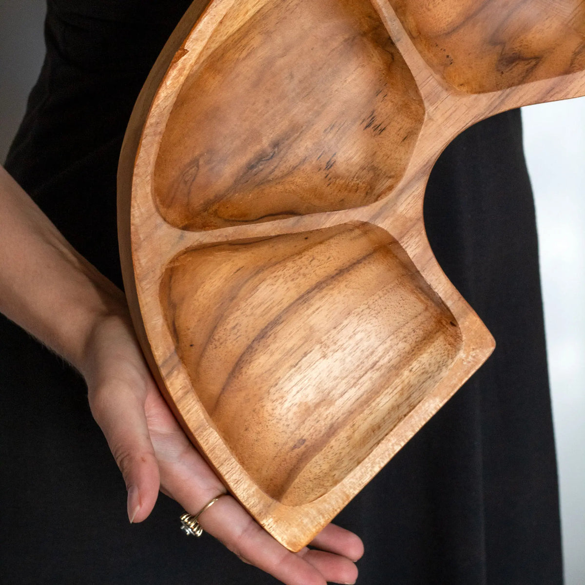 Cuenco de raíz de teca con forma curva y divisiones, sostenido por manos sobre fondo oscuro. Tono madera natural.