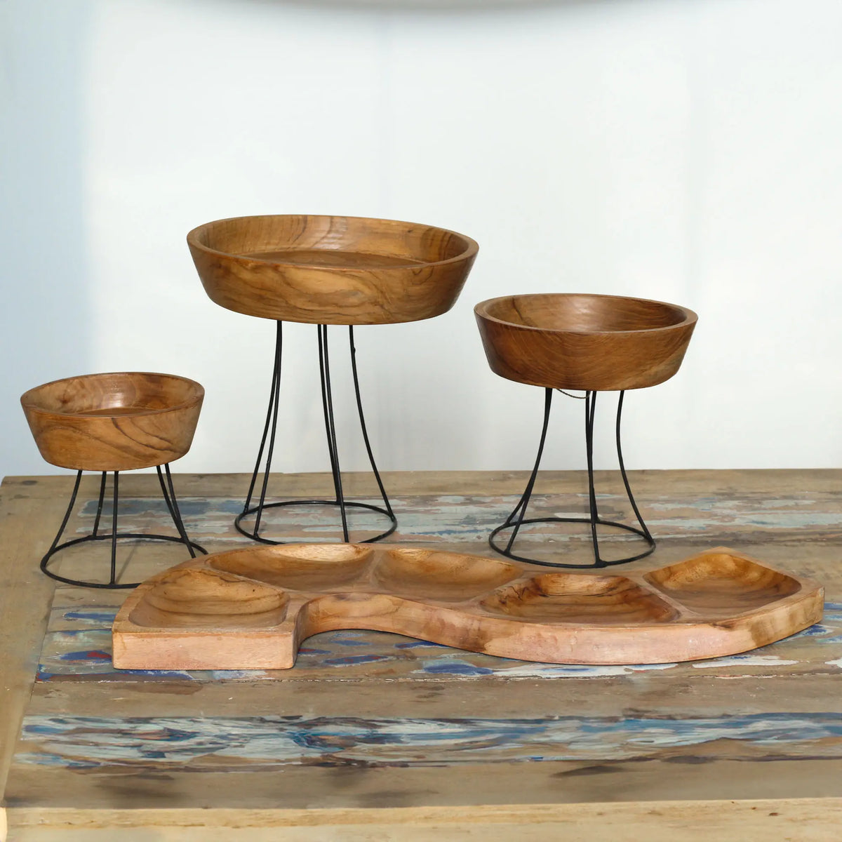 Cuenco alargado de madera de teca con forma de serpiente y tres cuencos elevados sobre bases de metal negro.