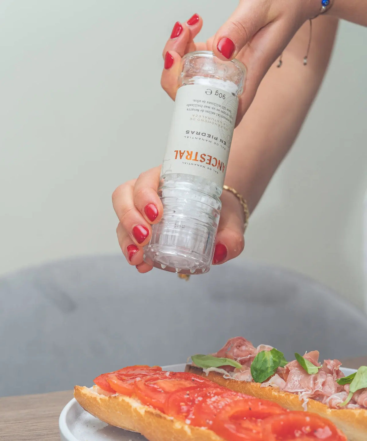 Hand holding a glass salt grinder over bruschetta, dispensing natural spring salt crystals onto tomato slices.