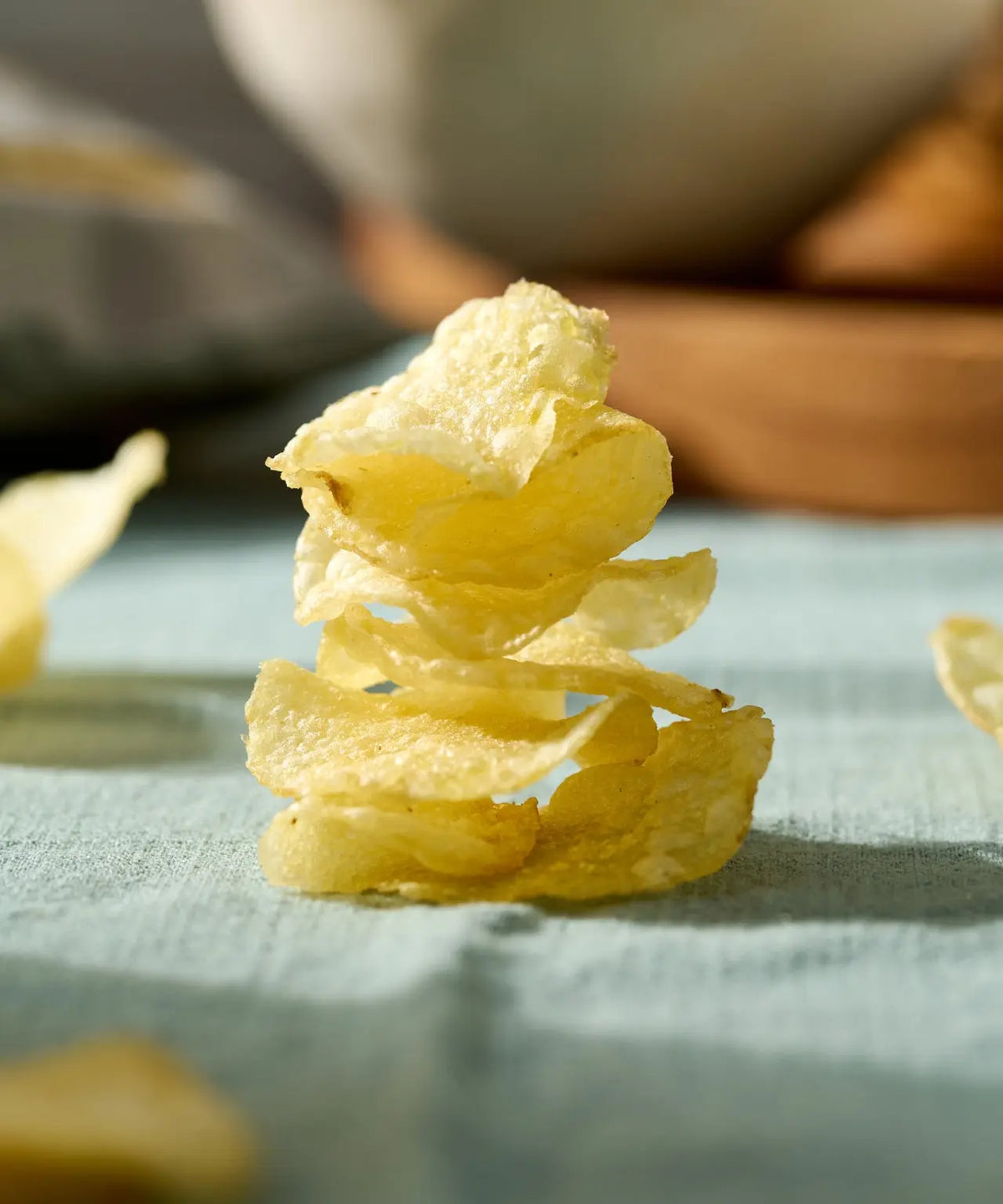 A vertical stack of golden, crispy potato chips on a textured, light blue fabric background in soft natural light.