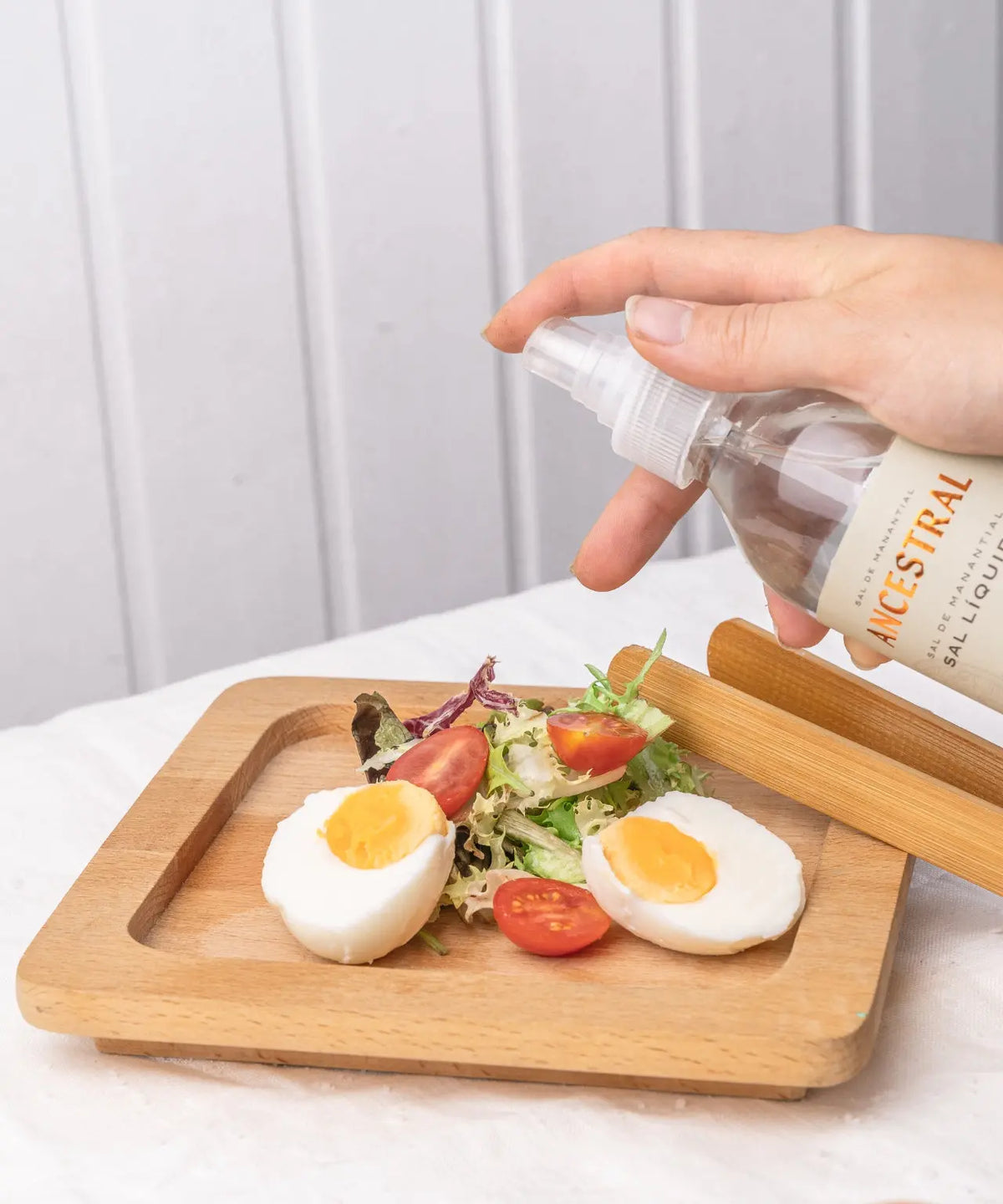 Hand spraying liquid salt from a 200ml bottle over a wooden board with halved boiled eggs, salad, and tomatoes.